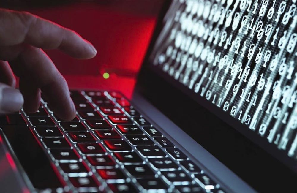 A man's fingers type on a laptop keyboard. There is computer code on the screen.