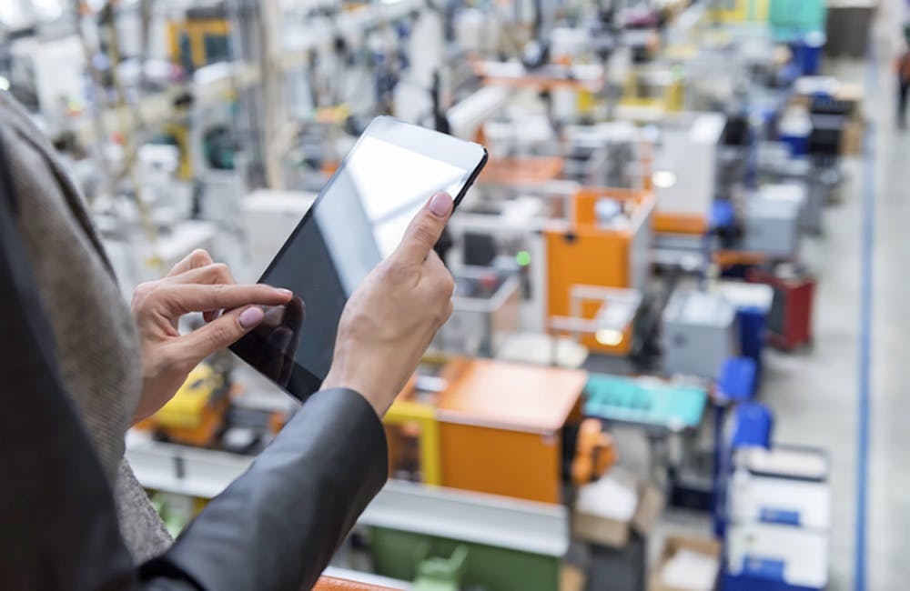 Horizontal color image of businesswoman - unrecognizable person - working with digital tablet in large futuristic factory. Woman standing on top of a balcony, holding touchpad and checking inventory of a manufacturing company on touchscreen tablet. Focus on businesswoman's hands holding black tablet, futuristic machines in background.