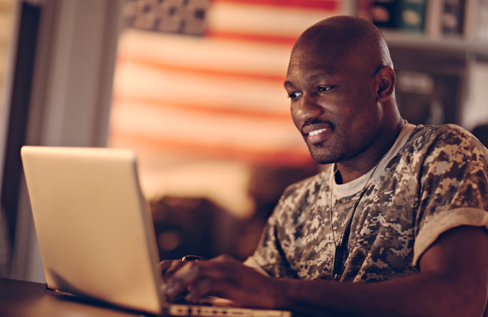 Picture of a young smiling african american male soldier sitting by a desk writing on his laptop. In the background is a bookshelf and on the wall is hanging an american flag. The soldier look tired but happy. From the left a dim light is sipping threw the window.