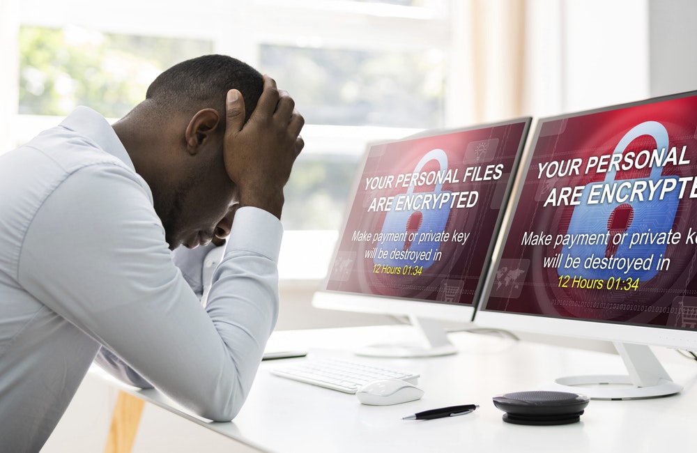 a man sits at desk with head in hands as computer screen reads "YOUR PERSONAL FILES ARE ENCRYPTED."