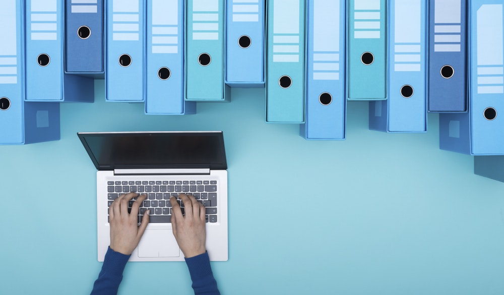 image of Organized archive with ring binders and woman searching for files in the database using a laptop, top view