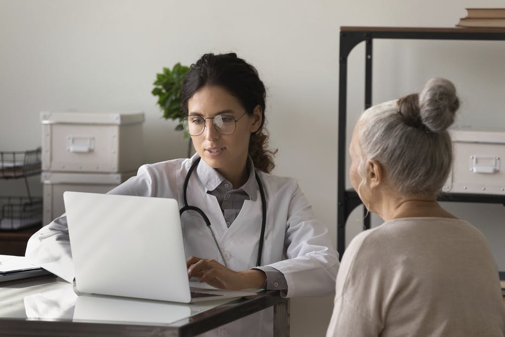 image of young female doctor meeting with elderly patient in office, listening to woman health problems complaints, typing on laptop, keeping electronic database on computer.