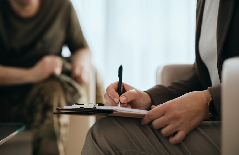 image of mental health professional writing notes while having meeting with a veteran.