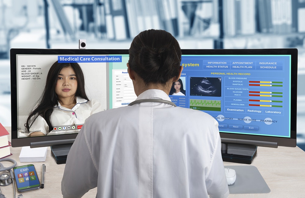 Female doctor working with two computers for distance patient consultation and searching her health information from medical record system.