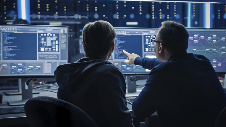 Two Professional IT Programers Discussing Blockchain Data Network Architecture Design and Development Shown on Desktop Computer Display. Working Data Center Technical Department with Server Racks