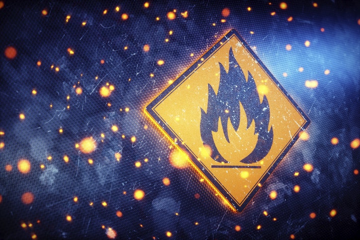 A glowing flame sign representing the international symbol for flammable materials or hazard. The sign is set on a dark blue scratched metallic wall, with glowing embers engulfing it.