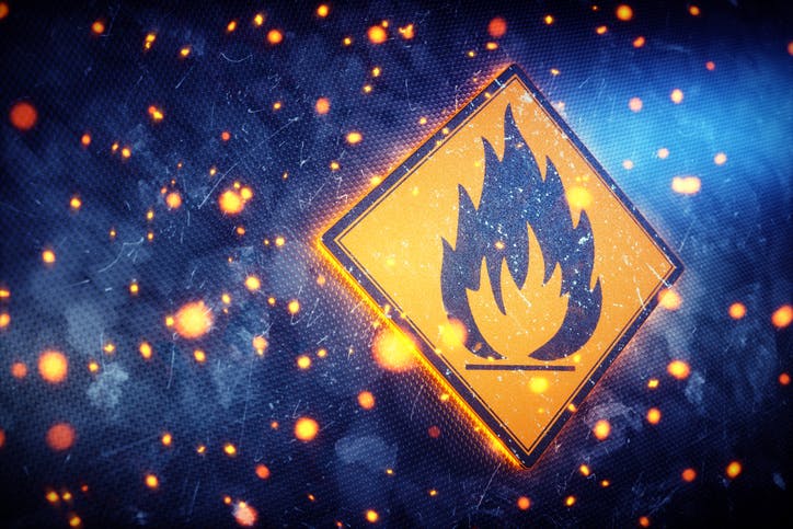 A glowing flame sign representing the international symbol for flammable materials or hazard. The sign is set on a dark blue scratched metallic wall, with glowing embers engulfing it.