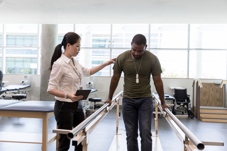 The female physical therapist guides the mid adult male military veteran as he walks on his foot during therapy.