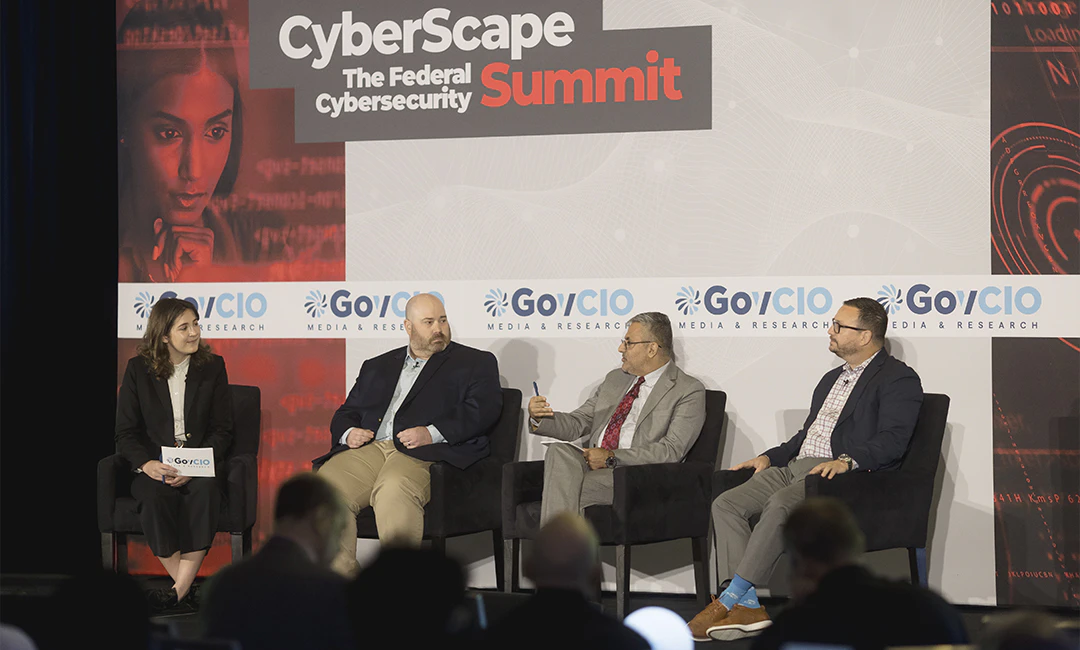 photo of the stage at GovCIO Media & Research's CyberScape: The Federal Cybersecurity Summit. Shows panelists sitting down in chairs on stage with Keith Busby, Acting CISO, CMS on the left, Anil Chaudhry, Senior Advisor, AI, Department of Transportation, in the center and Chris Saunders, WW Director of Public Sector Solutions Engineering, Wiz, on the right